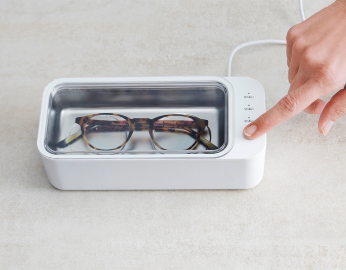 Brille in einem Ultraschallreiniger. Person drückt die 3-Minuten-Taste.