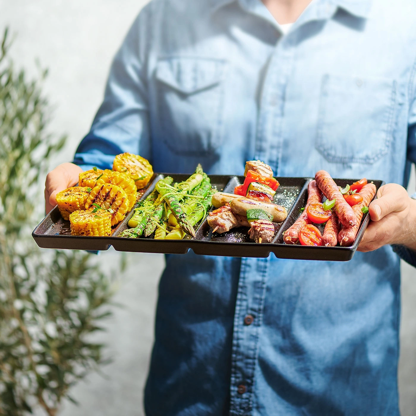 Person hält Grillteller mit Mais, Spargel, Würstchen und Gemüse