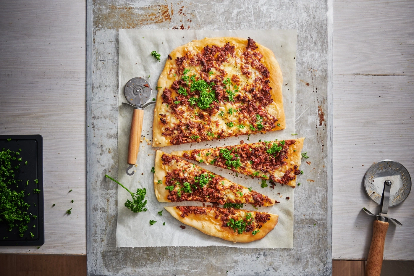 Pizza mit Fleisch und Petersilie, auf Papier, mit Pizzaschneider und Brettchen
