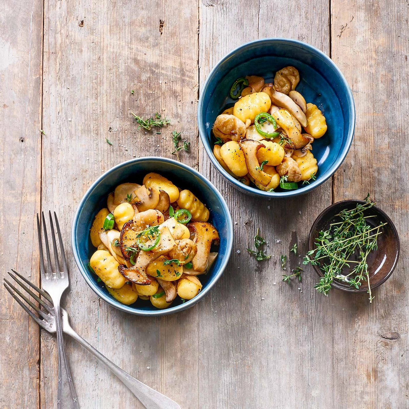 Zwei Schüsseln mit Pilz-Gnocchi auf einem Holztisch mit Kräutern und Gabeln.