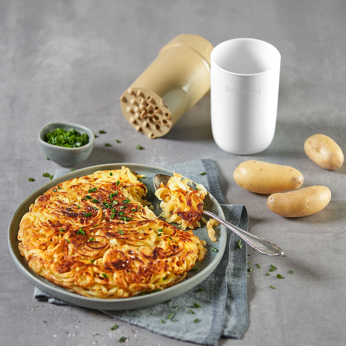 Kartoffelgericht auf Teller, mit Kräutern, daneben Kartoffeln und Utensilien