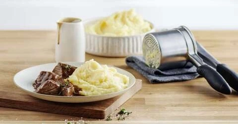 Teller mit Fleisch und Kartoffelpüree, daneben Soße und Kartoffelstampfer.
