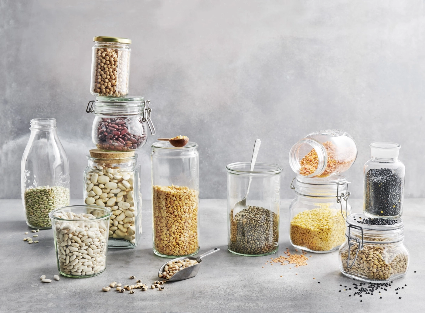 Des bocaux remplis de légumineuses, lentilles et haricots sur fond gris.