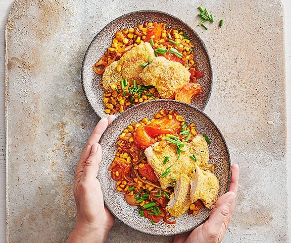 Zwei Teller mit gebratenem Hähnchen, Mais und Tomaten, von Händen gehalten.
