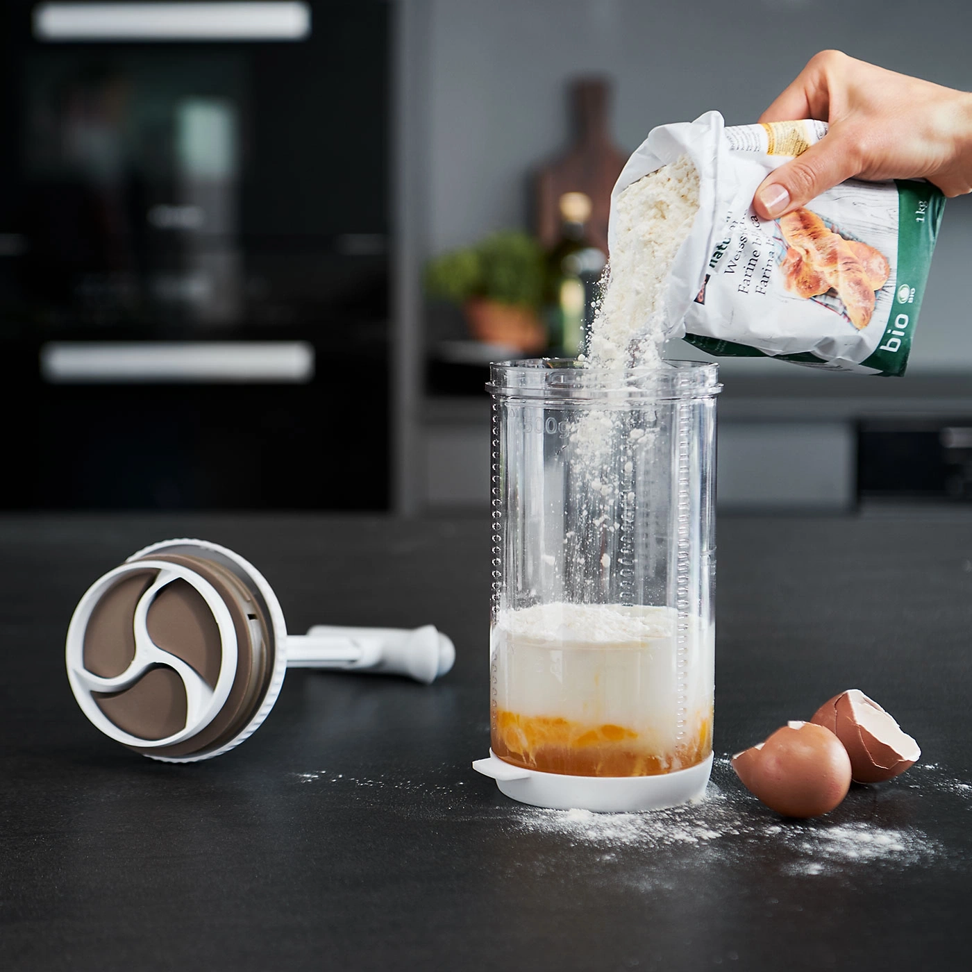 Mehl wird in ein Glas gegossen, Eier und ein Mixer liegen daneben.
