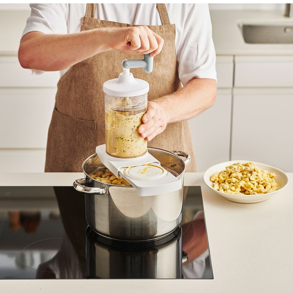 Une personne cuisine des spaetzle avec un appareil spécial dans la cuisine.
