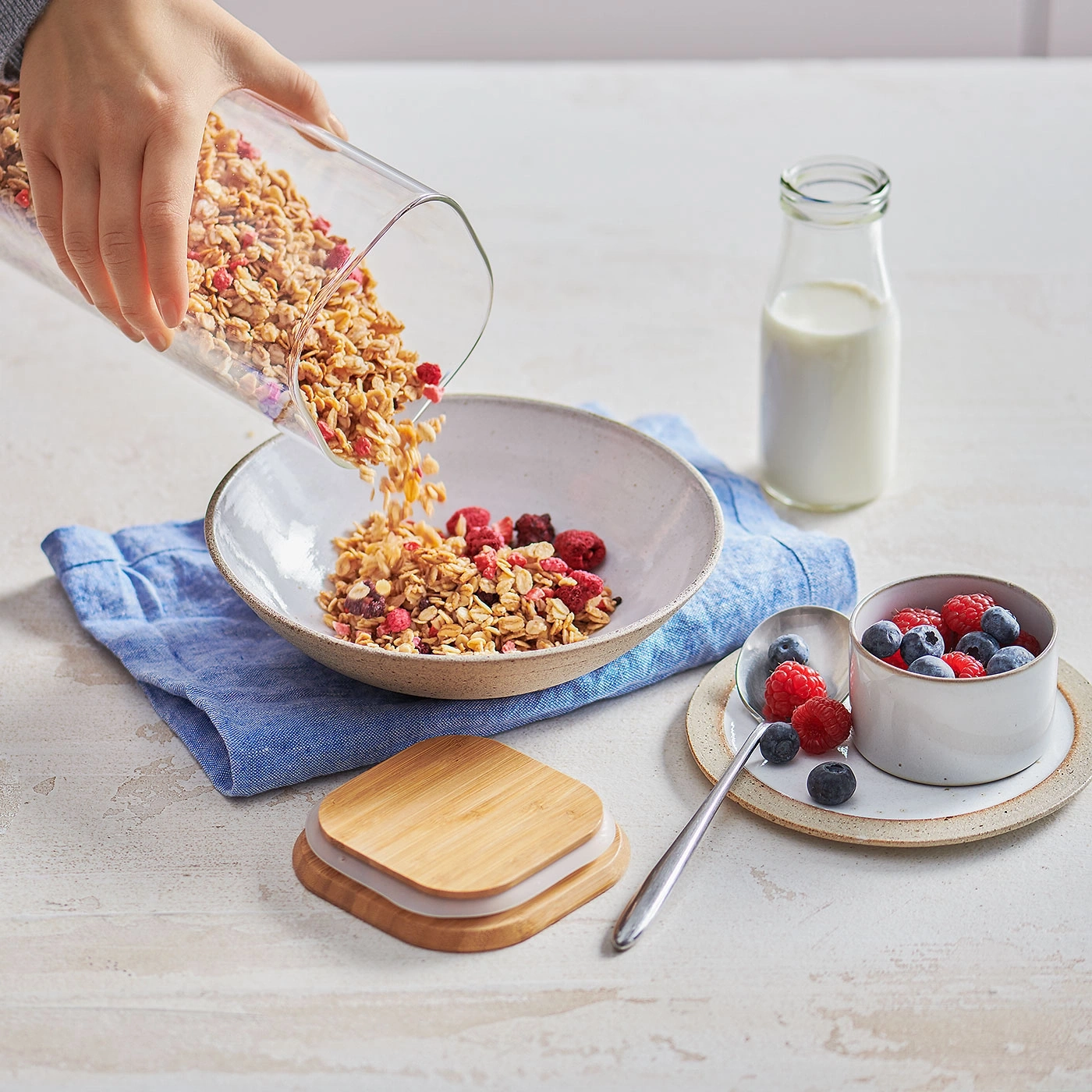 Person gießt Müsli in eine Schüssel mit Beeren und Milch daneben.
