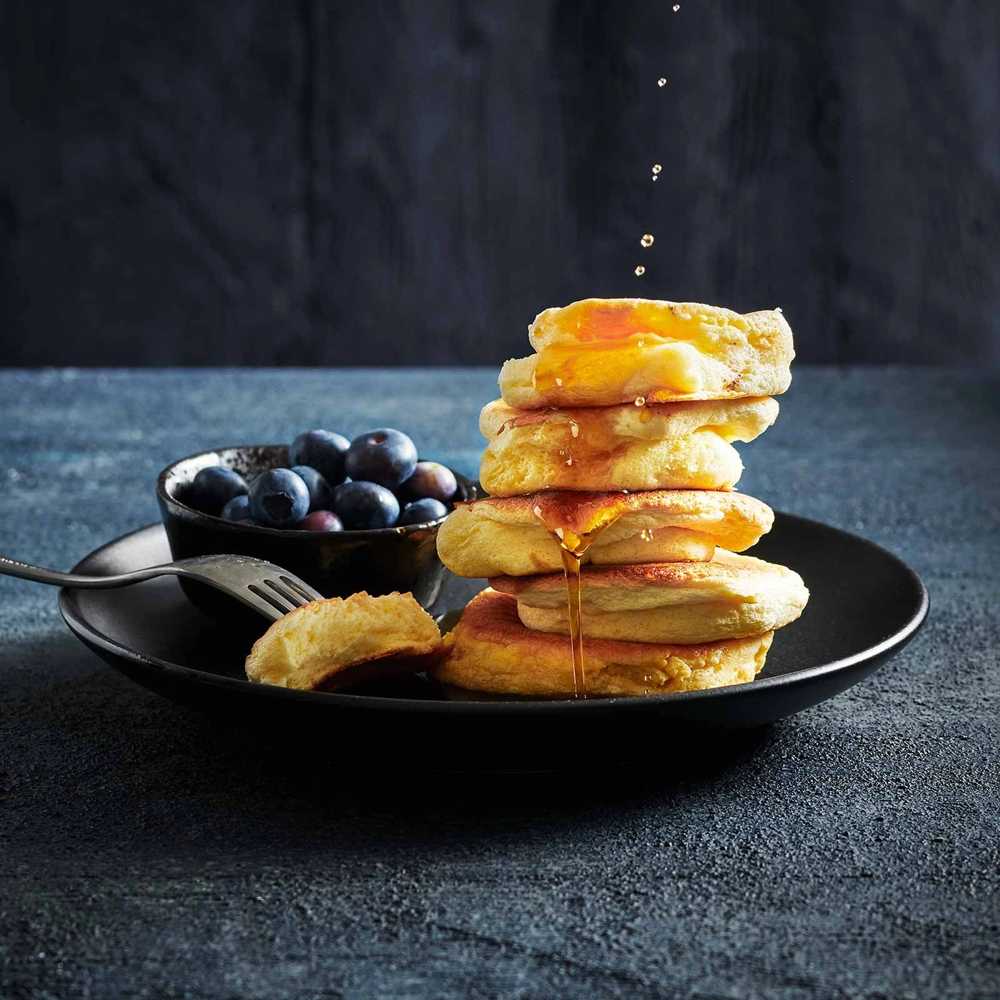 Stapel Pfannkuchen mit Blaubeeren und Honig, auf einem schwarzen Teller.