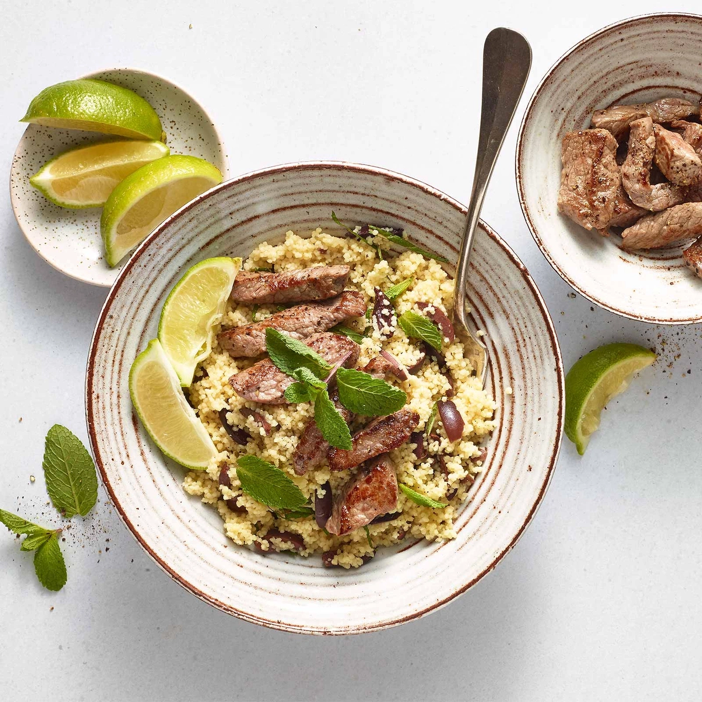 Couscous-Gericht mit Fleisch, Oliven, Limetten und Minze in Schalen auf weißem Hintergrund.
