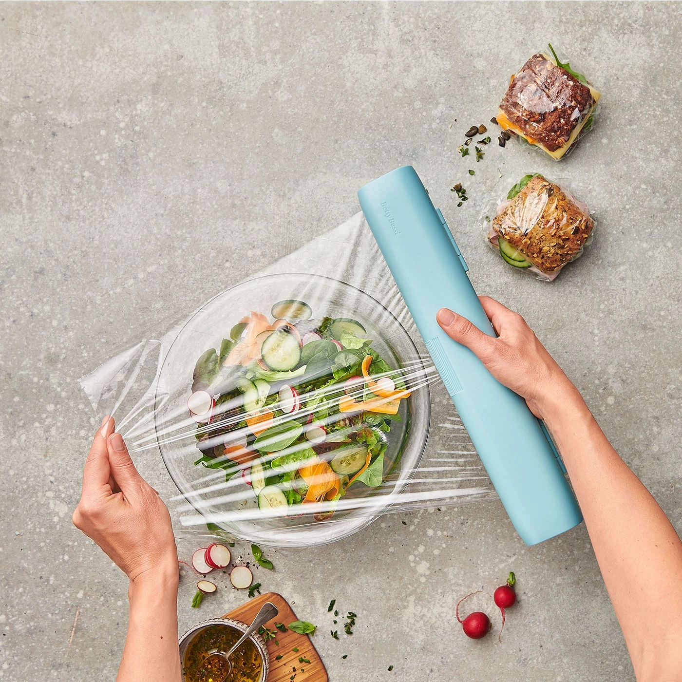 Salat mit Frischhaltefolie verpackt, Sandwich, blaues Werkzeug, grauer Hintergrund.