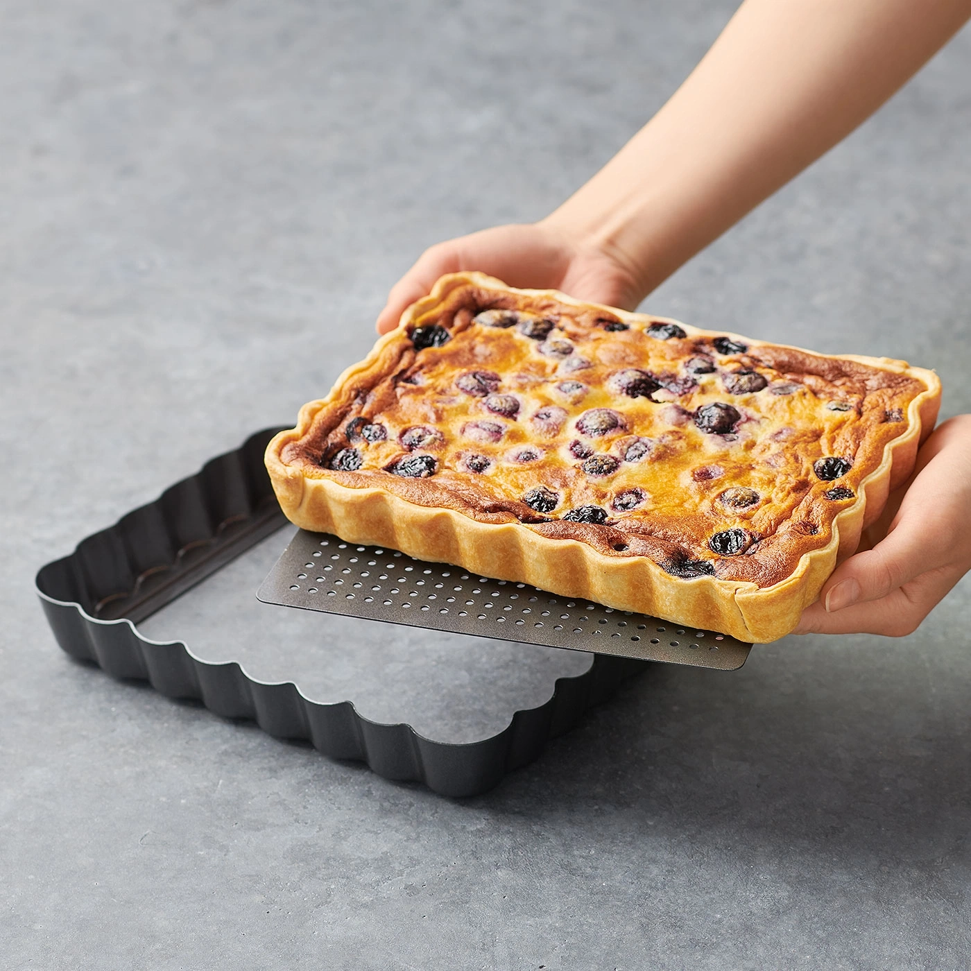 Person hält Blaubeerkuchen aus rechteckiger Backform. Kuchen wird herausgenommen.