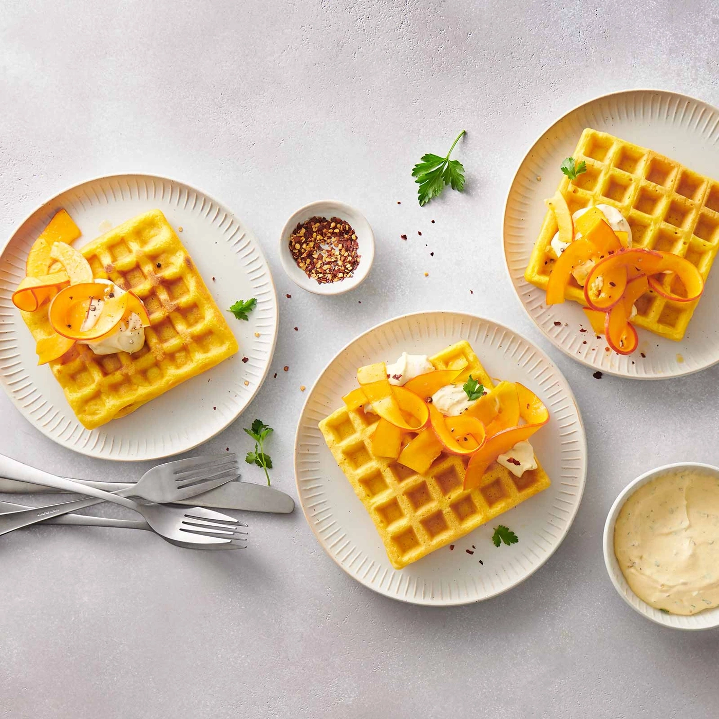 Waffeln mit Belag, Chili, Kräutern und Soße auf Tellern, Gabeln und Messer.