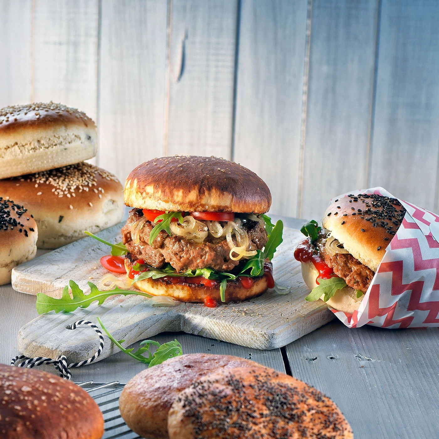 Burger mit Tomaten, Zwiebeln und Rucola auf Holzbrett, Brötchen im Hintergrund