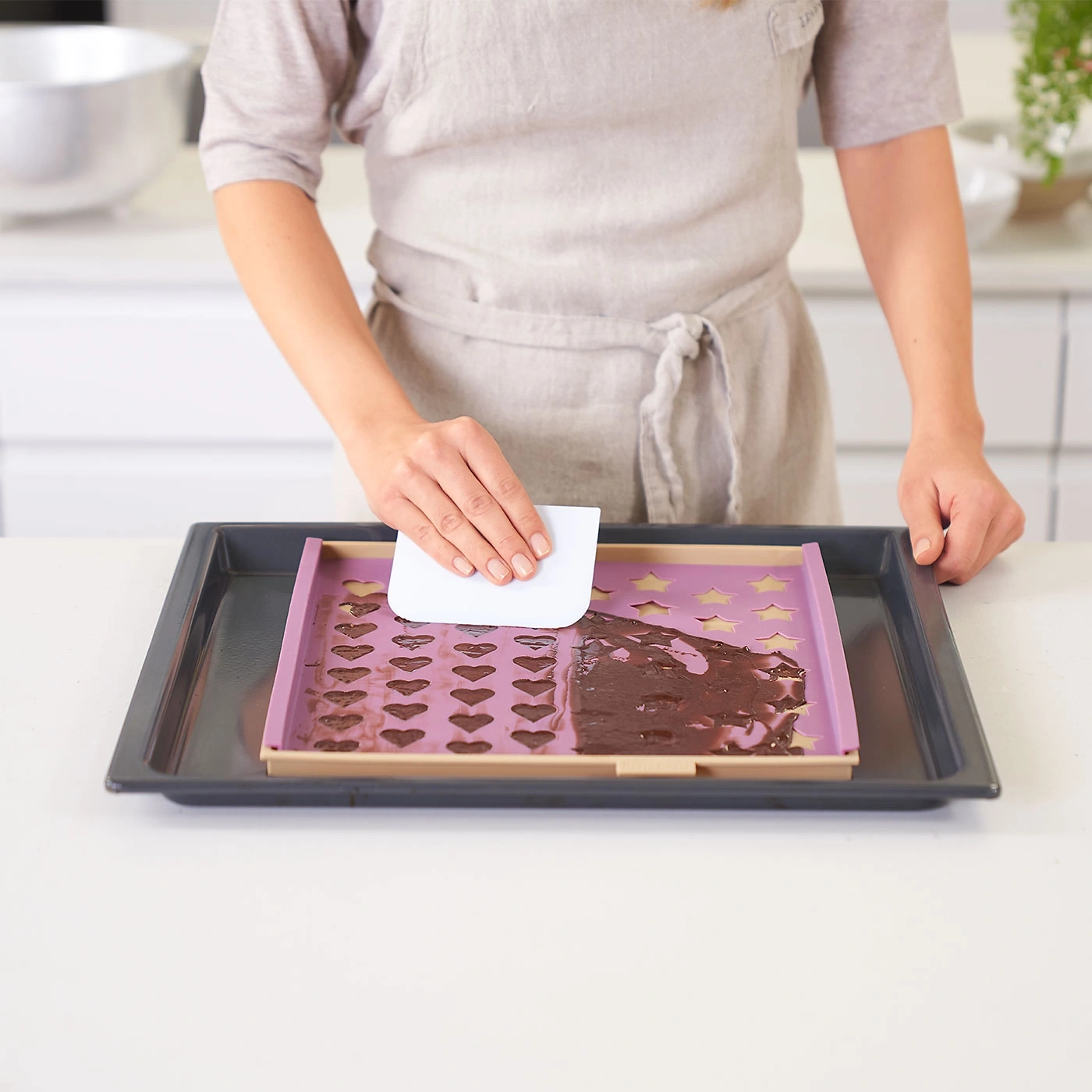 Personne en tablier étale du chocolat avec une spatule sur un moule à pâtisserie avec des étoiles et des coeurs.