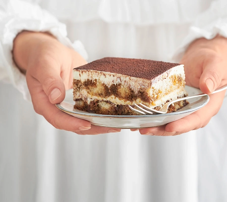 Person hält ein Stück Tiramisu auf einem Teller mit einer Gabel.