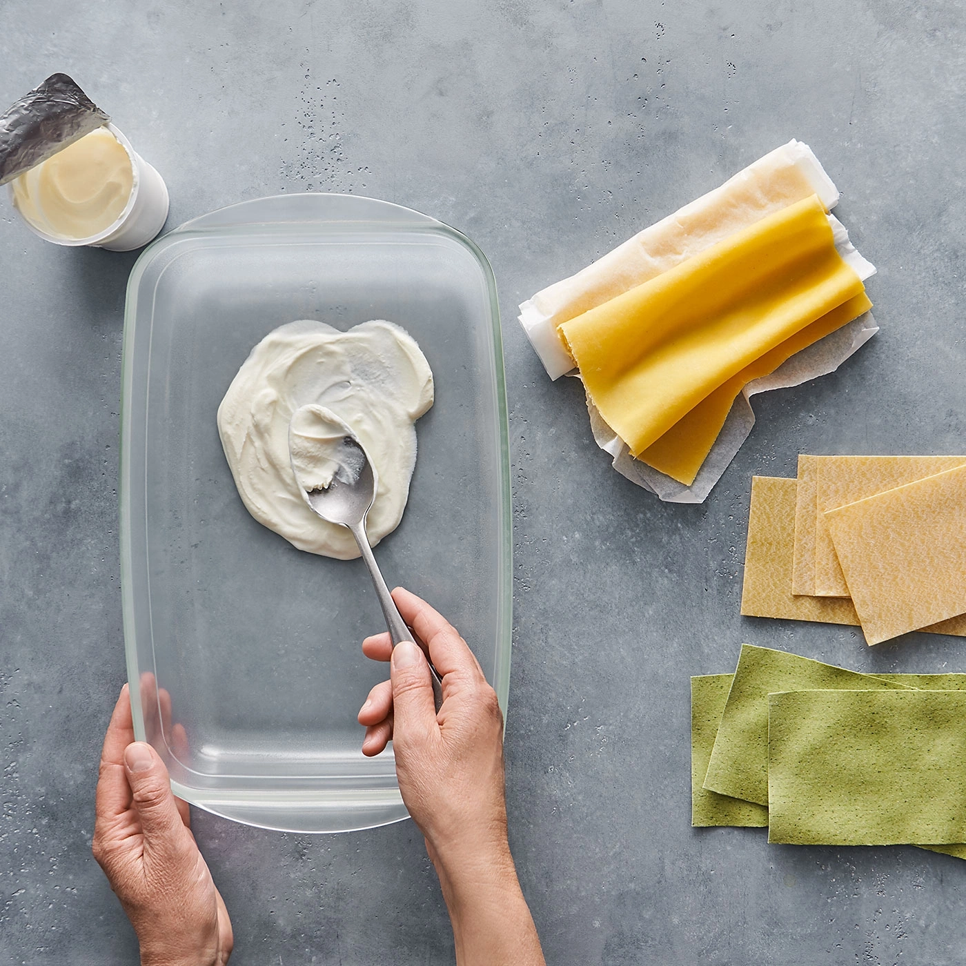 Lasagne-Zutaten: Nudeln, Käse, Schüssel, Löffel, Hände auf grauem Hintergrund.