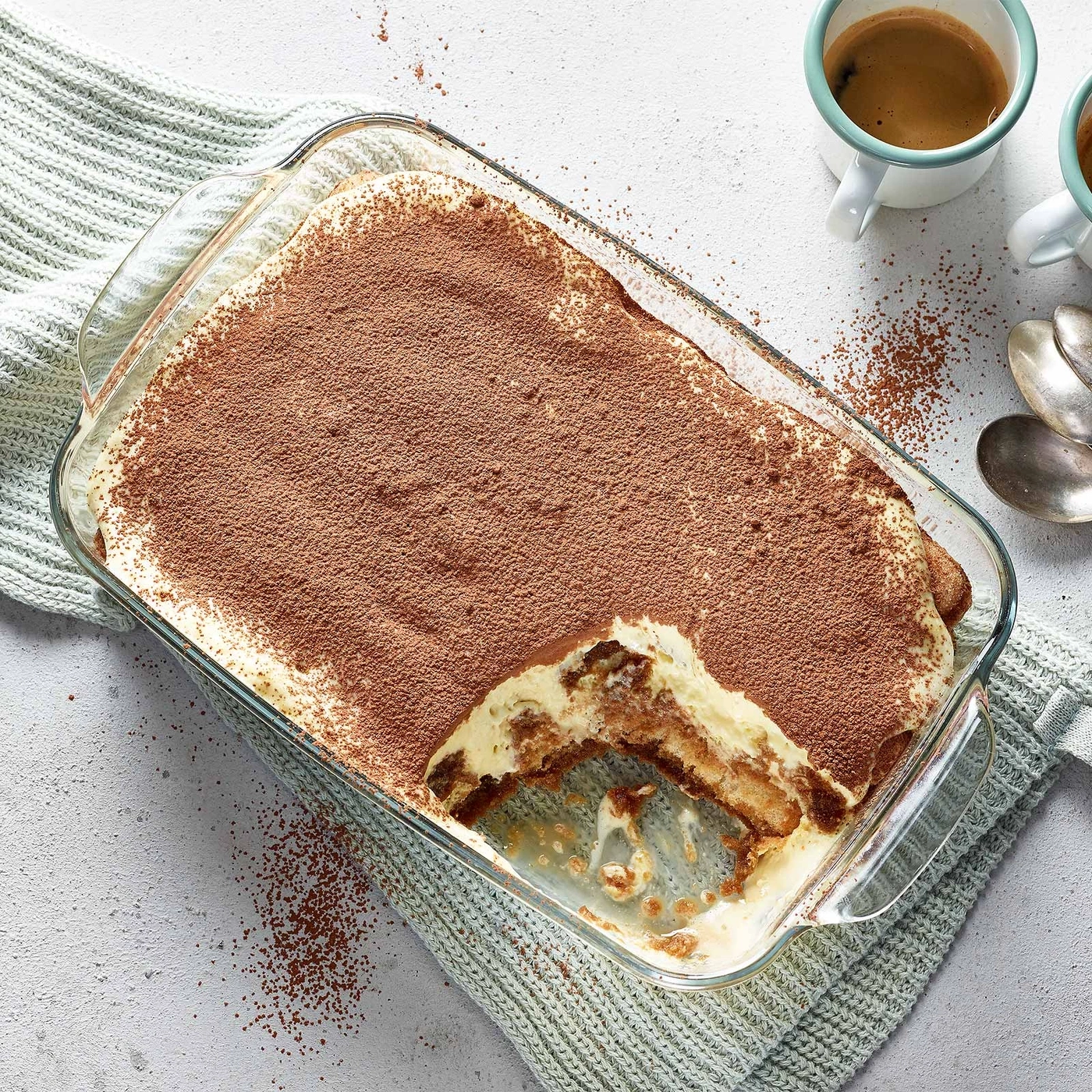 Tiramisu in Glasform, mit Kakaopulver bestreut, daneben Kaffee und Löffel