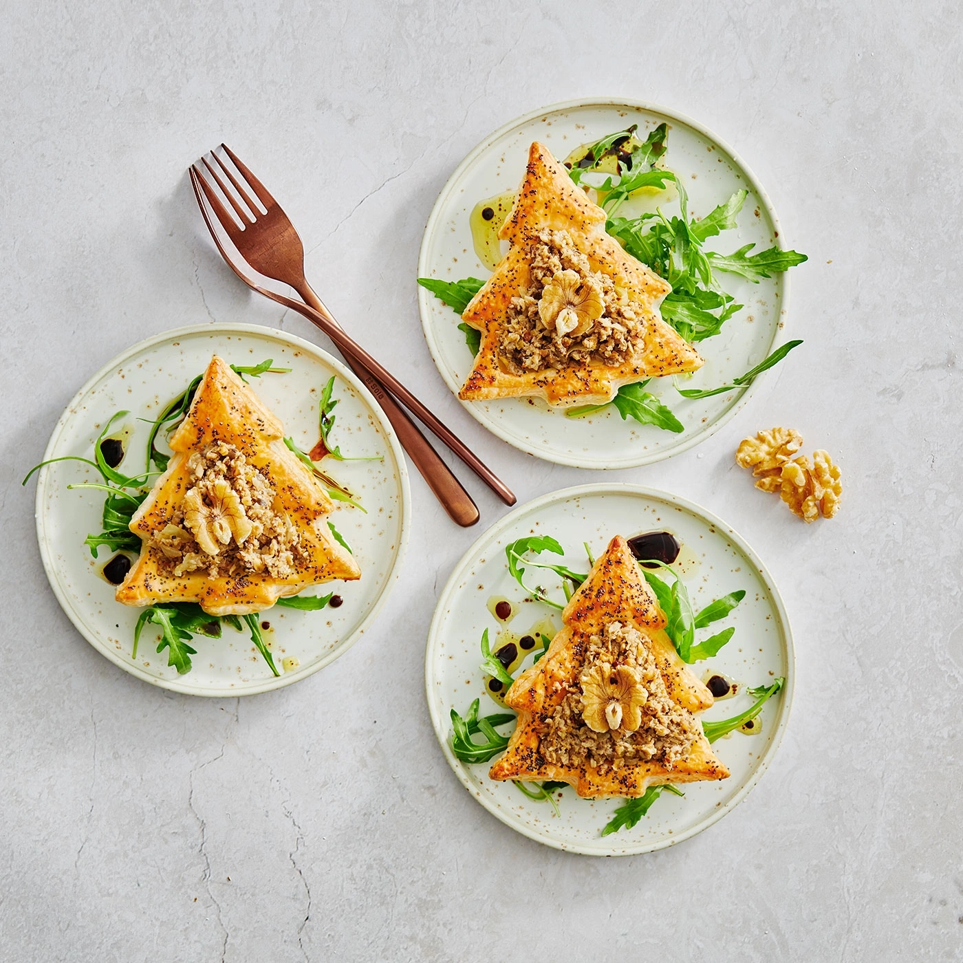 Trois assiettes avec pâtisseries en forme de sapin, roquette et noix sur fond clair.