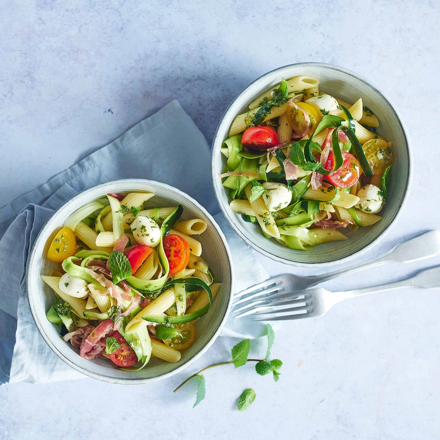 Zwei Schüsseln Pasta-Salat mit Tomaten, Mozzarella, Zucchini und Minze.