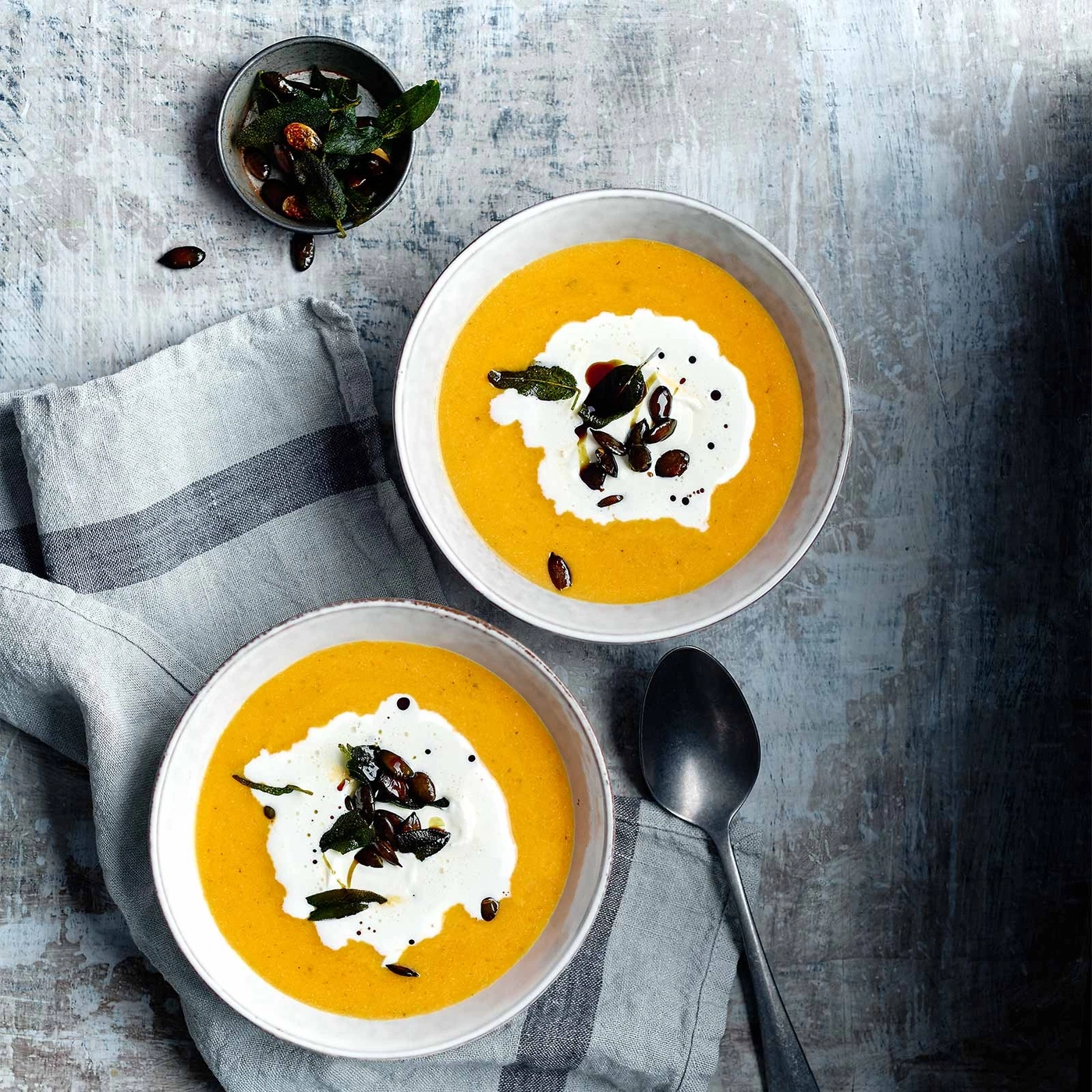 Zwei Schüsseln Kürbissuppe mit Sahne, Salbei und Samen auf einem Tisch.