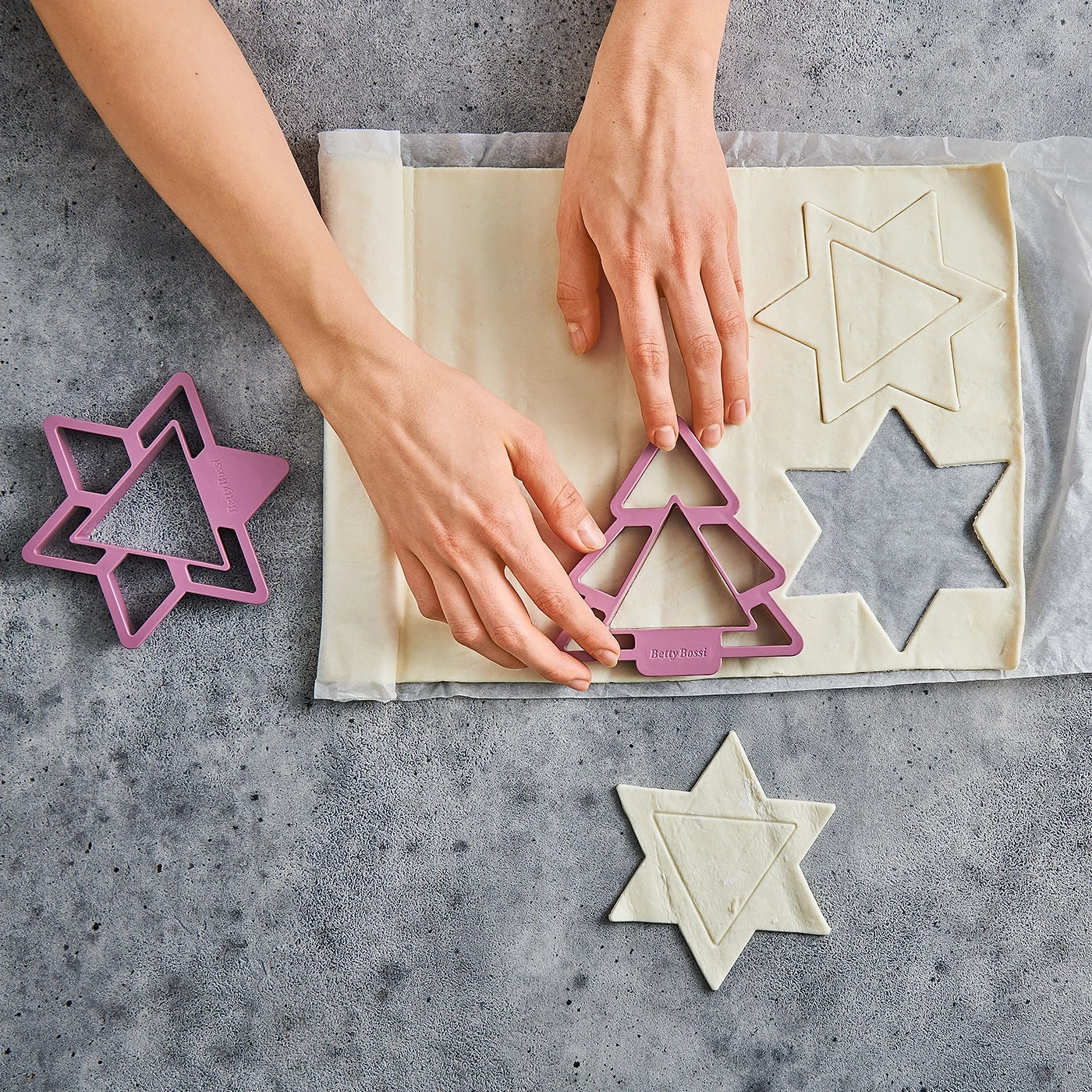 Mains coupant la pâte avec des emporte-pièces en forme d'étoile et de sapin sur fond gris.