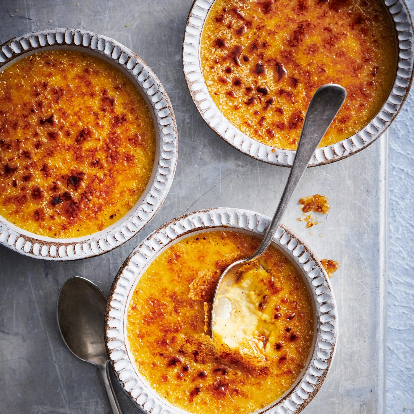 Drei Crème brûlée in Schalen mit Löffeln auf einem Tablett. Köstlich.