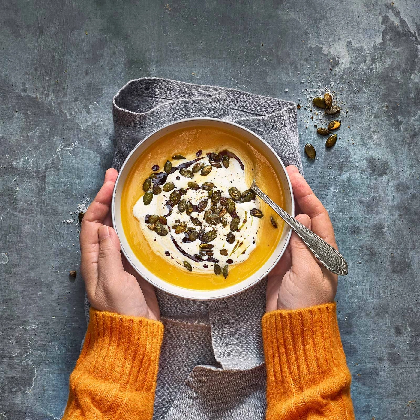 Kürbissuppe mit Sahne, Samen und Balsamico-Glasur in einer Schüssel, gehalten von Händen.