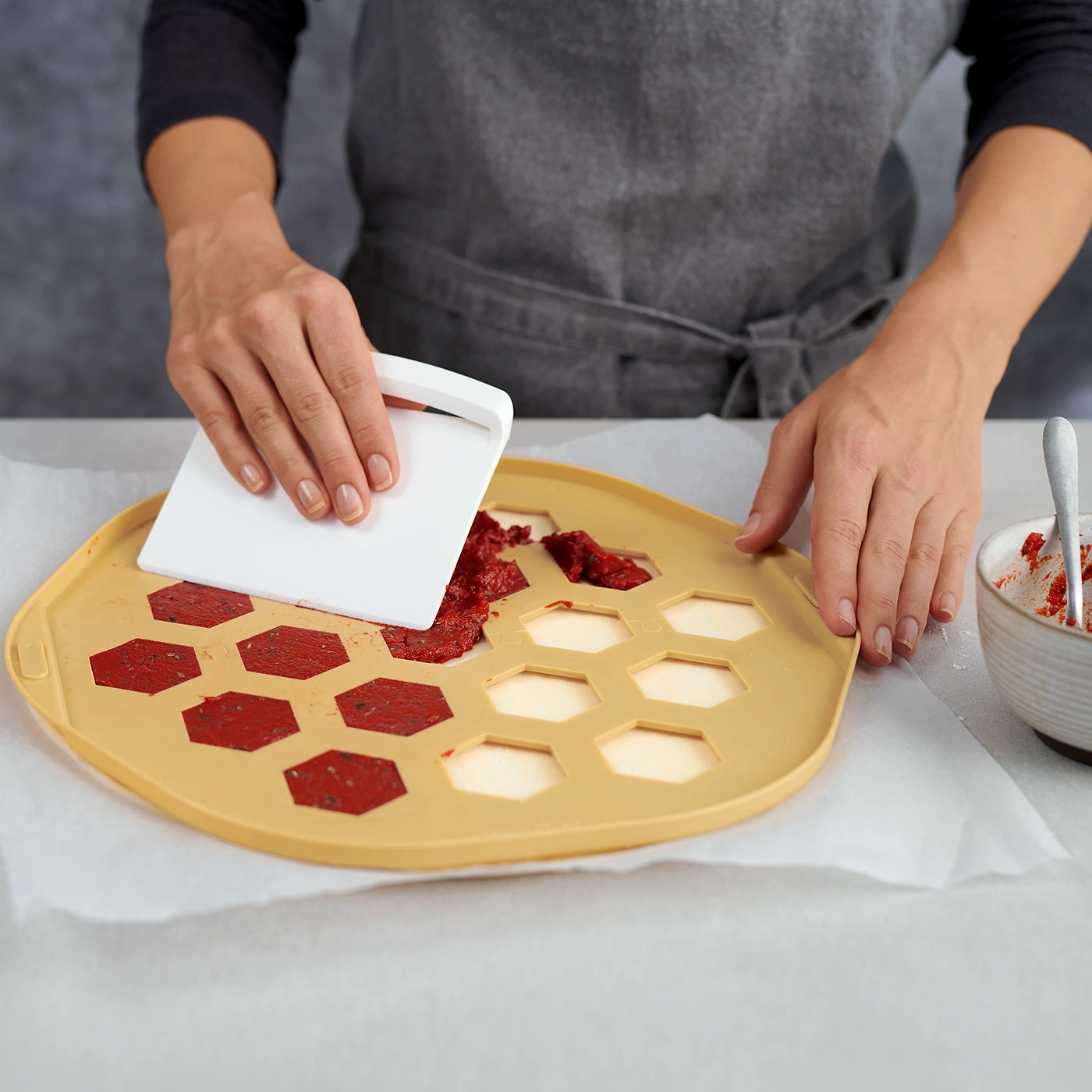 Une personne utilise une spatule pour étaler une pâte rouge sur un plateau jaune.