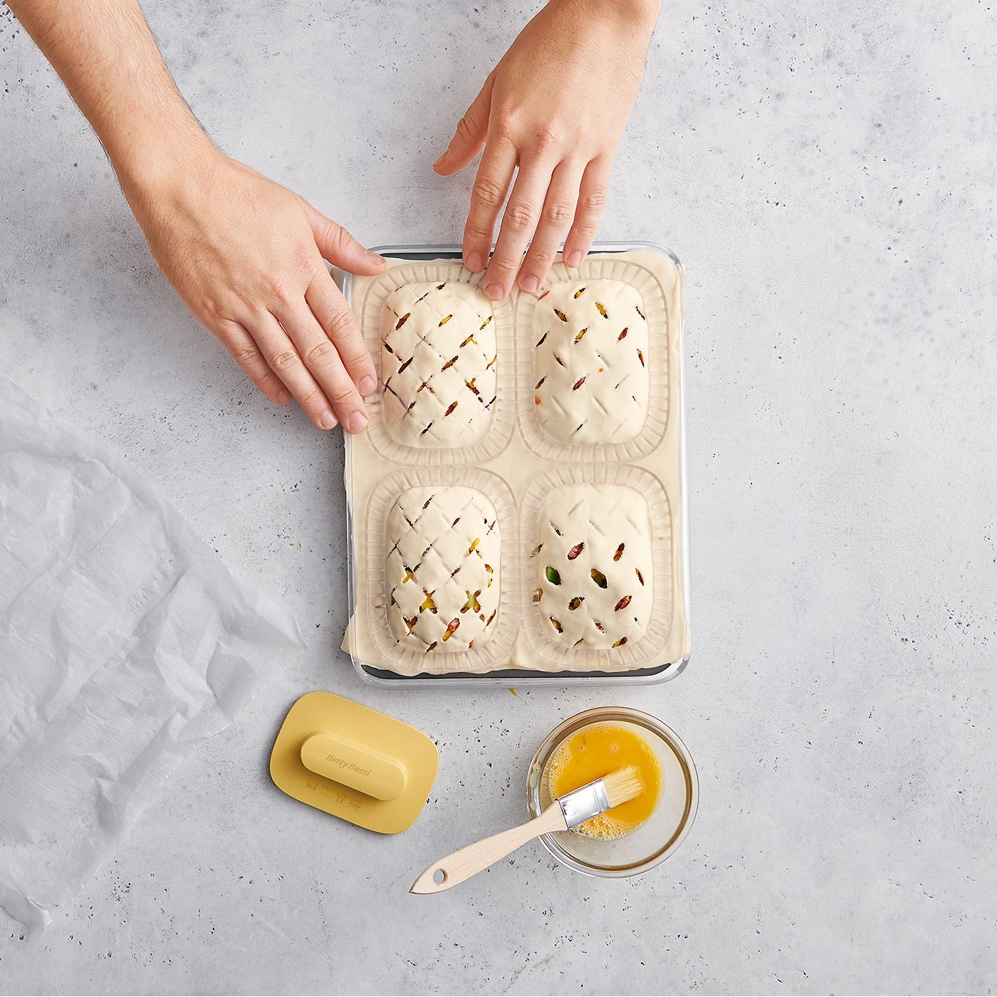 Mains façonnant des pâtisseries, pinceau et jaune d'œuf sur fond gris, préparation culinaire.