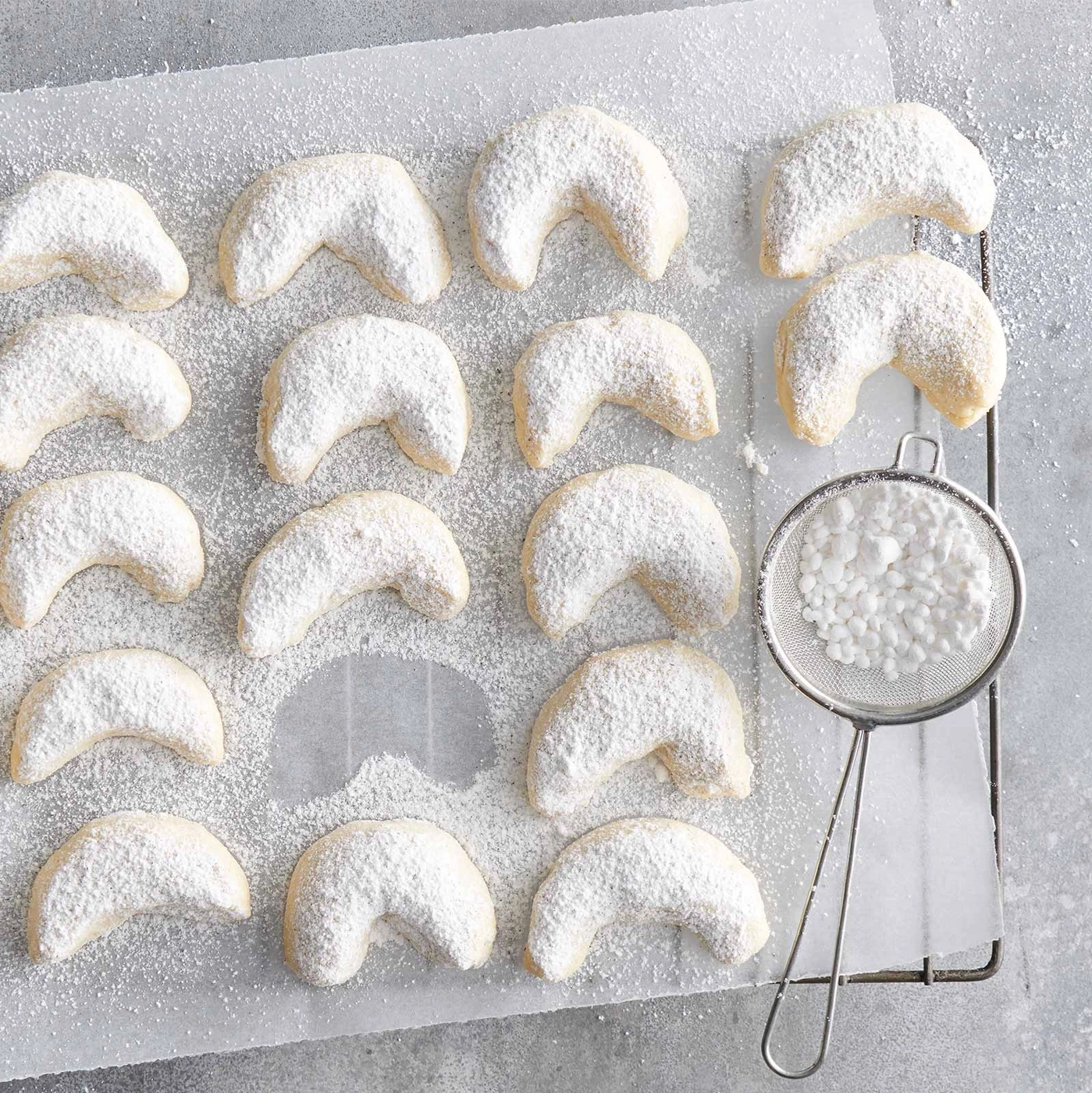 Vanillekipferl auf Backpapier, mit Puderzucker bestäubt, daneben ein Sieb.