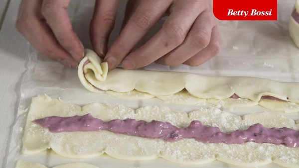Mains roulant une pâtisserie avec une garniture aux myrtilles, logo Betty Bossi en arrière-plan.