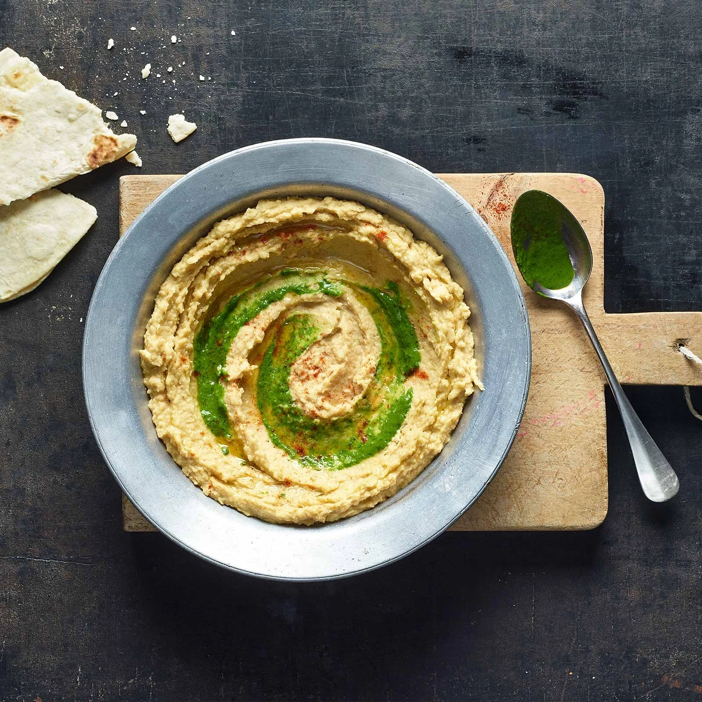 Hummus mit Kräuteröl, Fladenbrot und Löffel auf Holzbrett