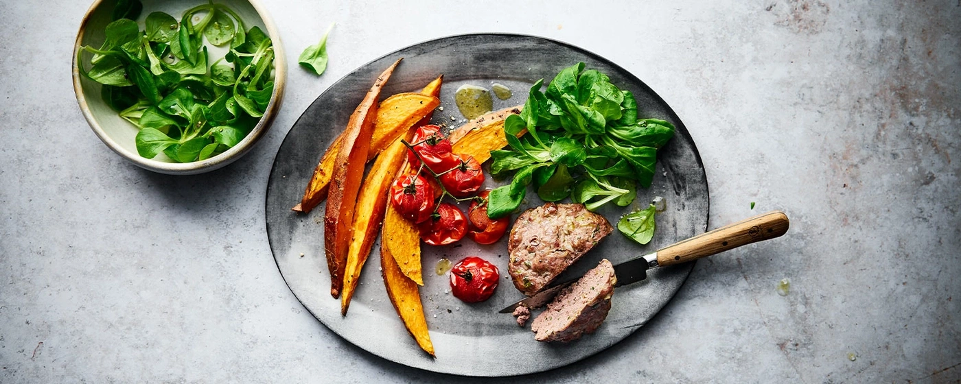 Teller mit Fleisch, Gemüse und Salat, serviert auf einem grauen Untergrund.