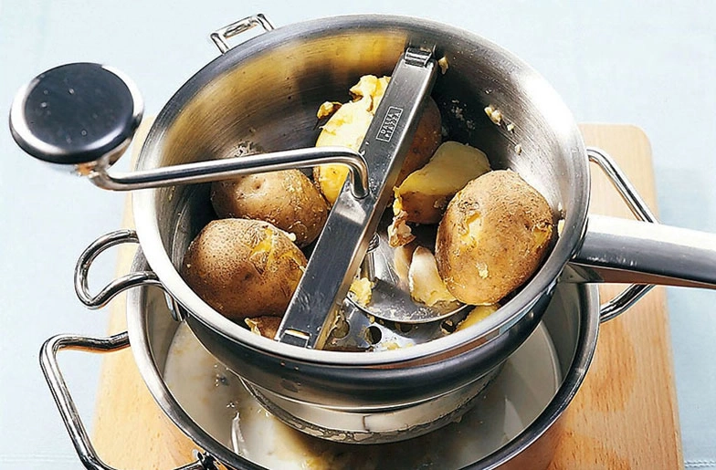 Kartoffeln in einem Topf mit Kartoffelpresse auf Holzbrett.