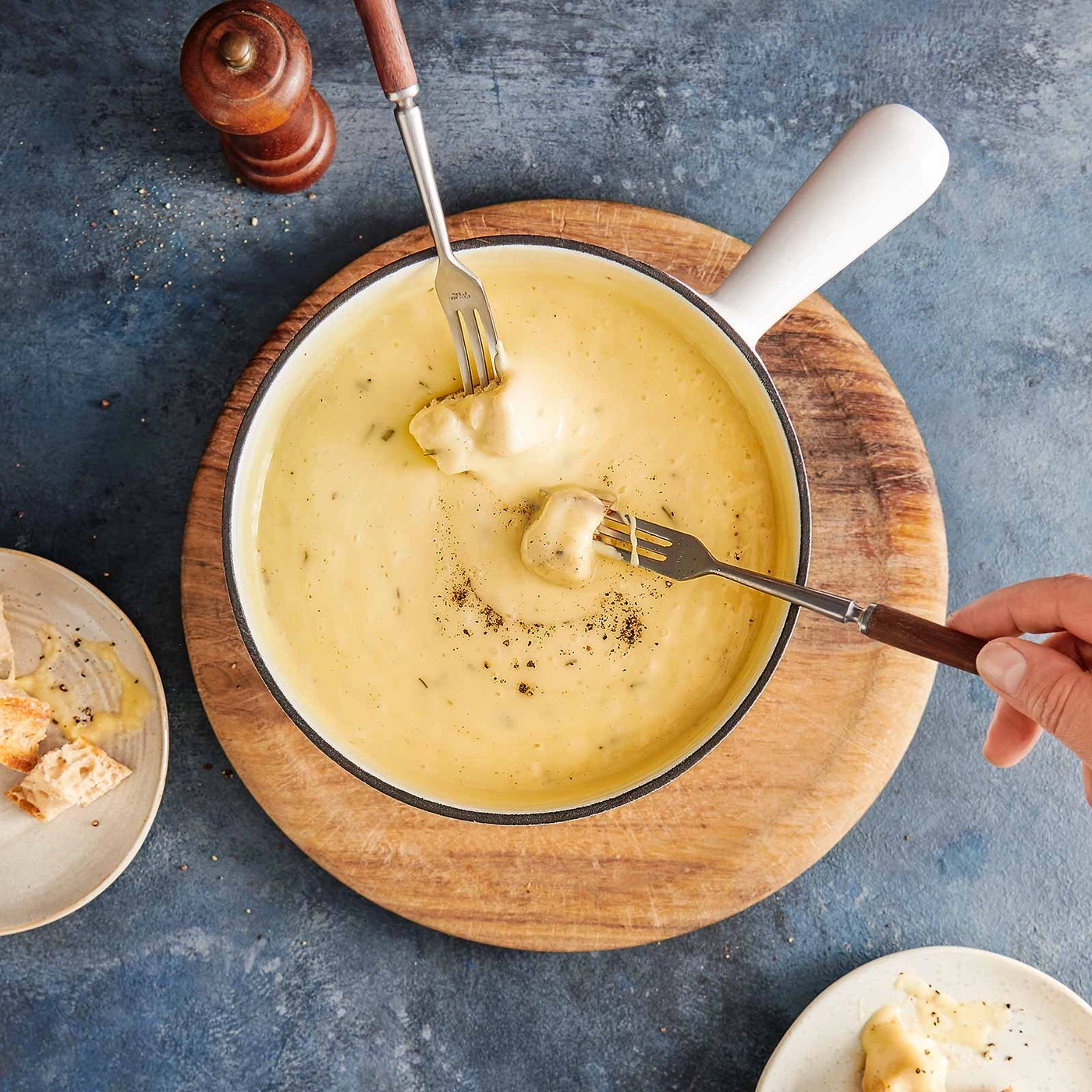Käsefondue mit Brot auf einem Holzbrett, Gabeln und Pfefferstreuer.
