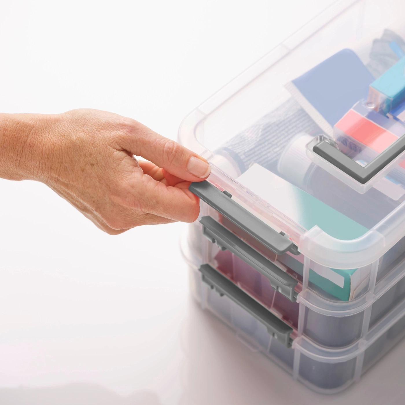 Hand öffnet eine transparente Aufbewahrungsbox mit grauem Verschluss, Inhalt sichtbar.
