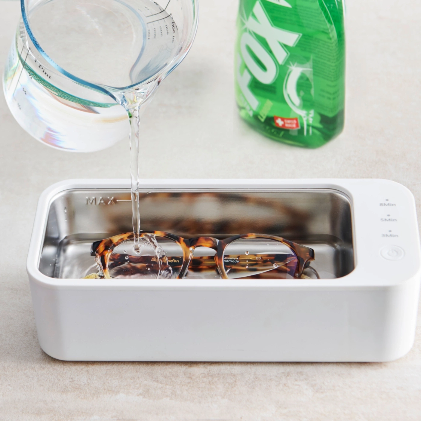Des lunettes sont nettoyées dans un nettoyeur à ultrasons avec de l'eau, à côté du liquide vaisselle.
