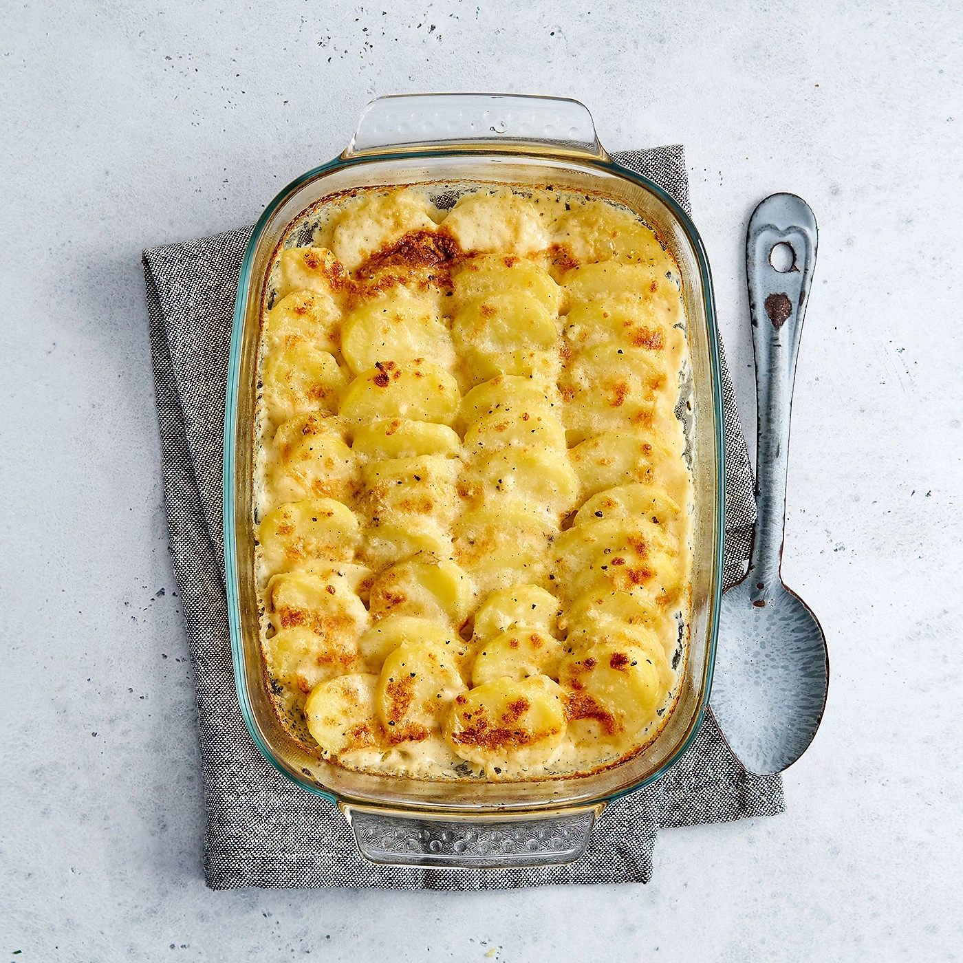 Kartoffelauflauf in Glasform mit Löffel auf grauem Tuch