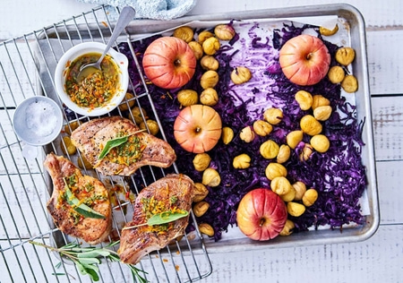 Gebratene Koteletts mit Äpfeln, Kastanien und Rotkohl auf einem Backblech.