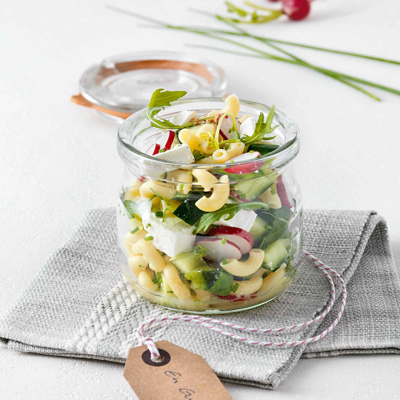 Glas mit Salat, Nudeln, Gemüse und Käse auf einem grauen Tuch.