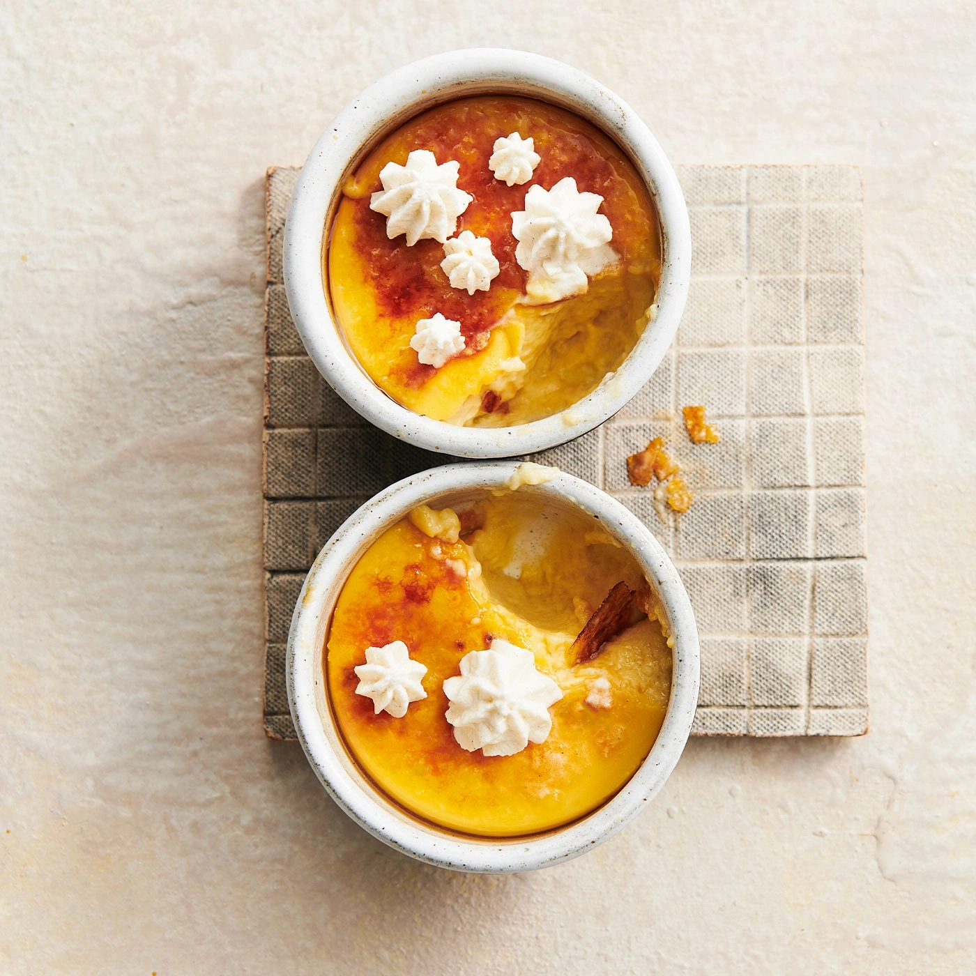 Zwei Crème brûlée in Schalen mit Sahne dekoriert, auf einem quadratischen Untersetzer.